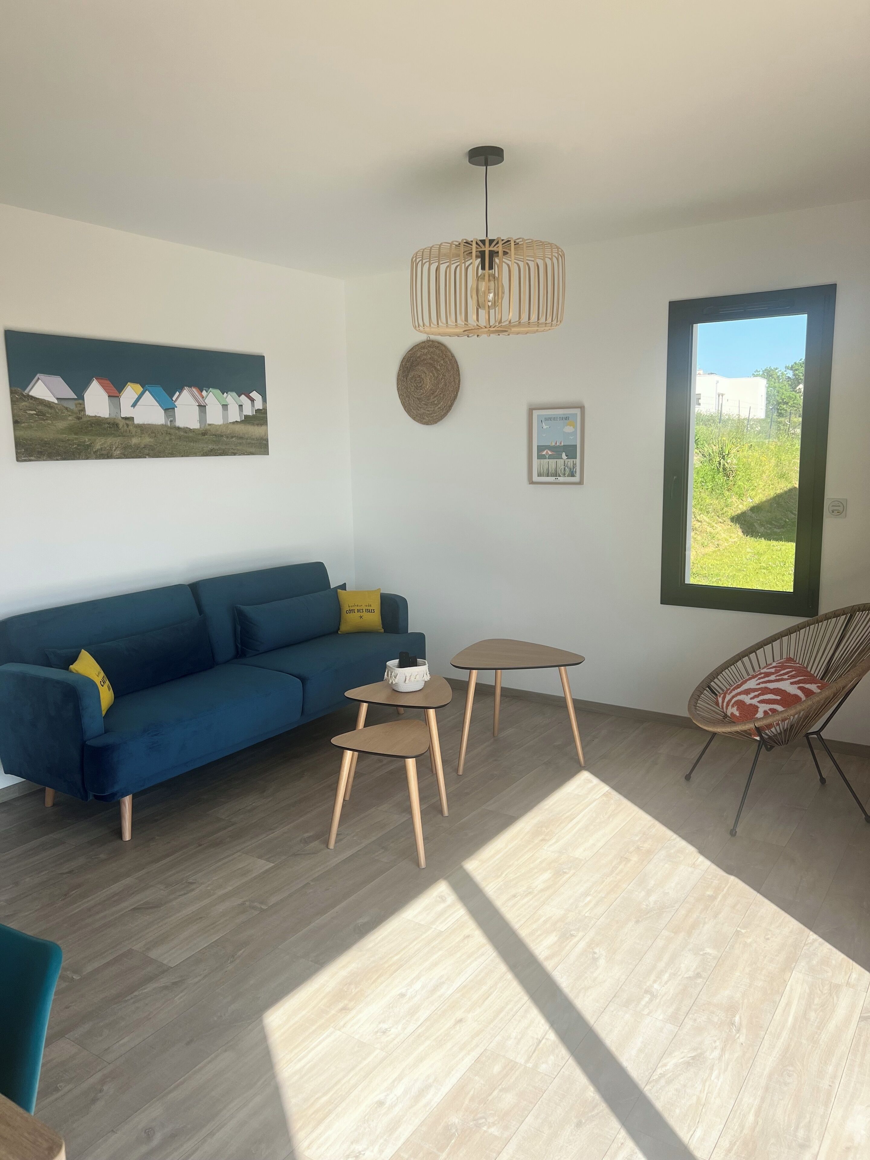Un salon moderne avec un canapé bleu, une table basse ronde, une petite table d’appoint, un fauteuil en osier et une grande fenêtre donnant sur un paysage verdoyant.