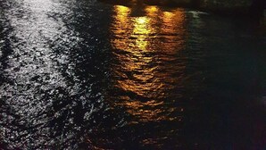 Escena costera nocturna con cielo iluminado por la luna, mar tranquilo que refleja luces y una costa distante iluminada.