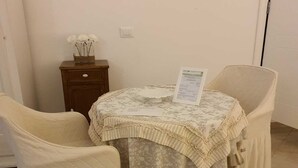 Un rincón comedor con dos sillas, una mesa con mantel floral, una mesita de madera con un jarrón de flores blancas y un interruptor de luz en la pared.