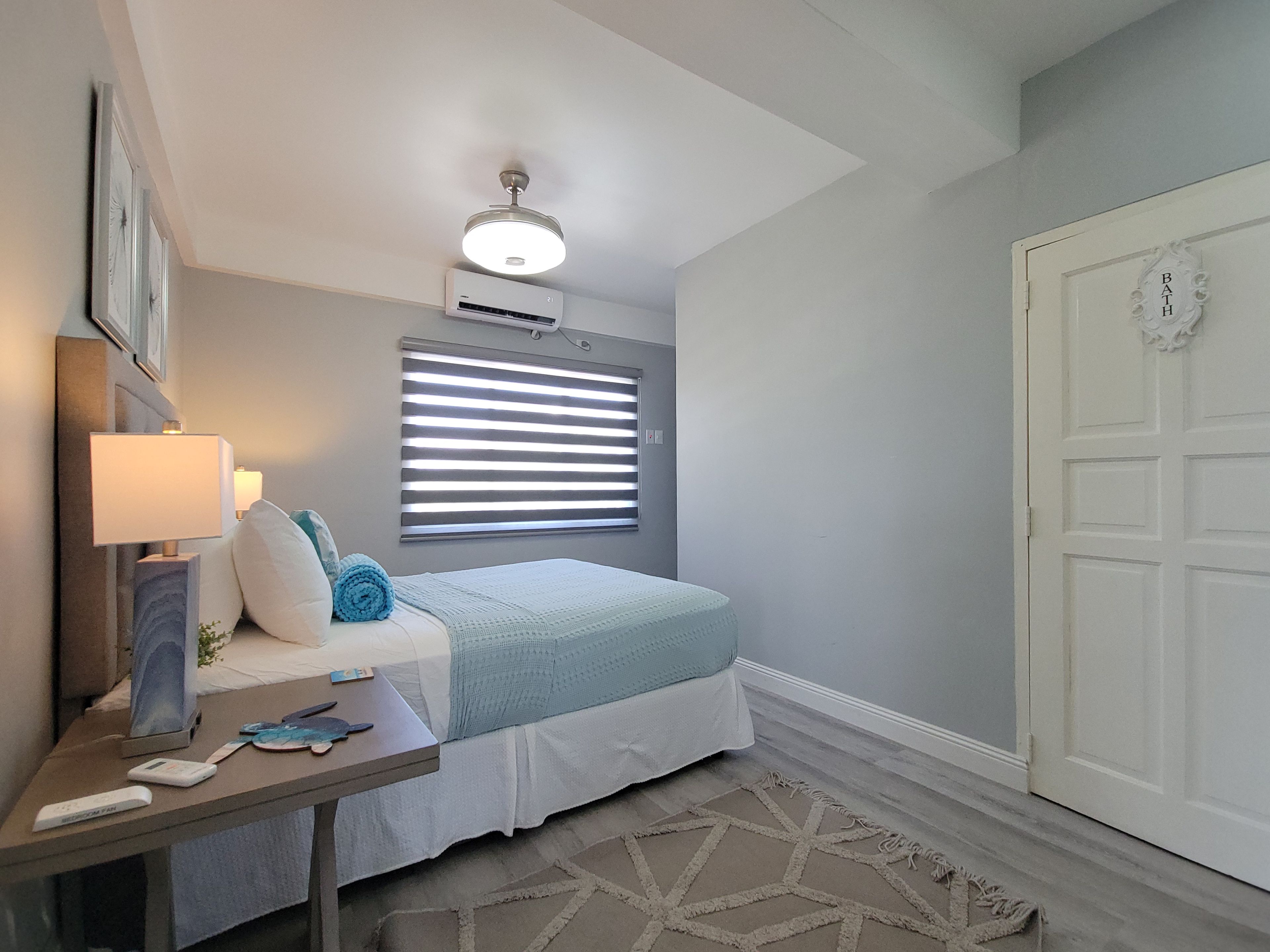 A bedroom with a bed, bedside table, lamp, air conditioning unit, window with blinds, and a door.