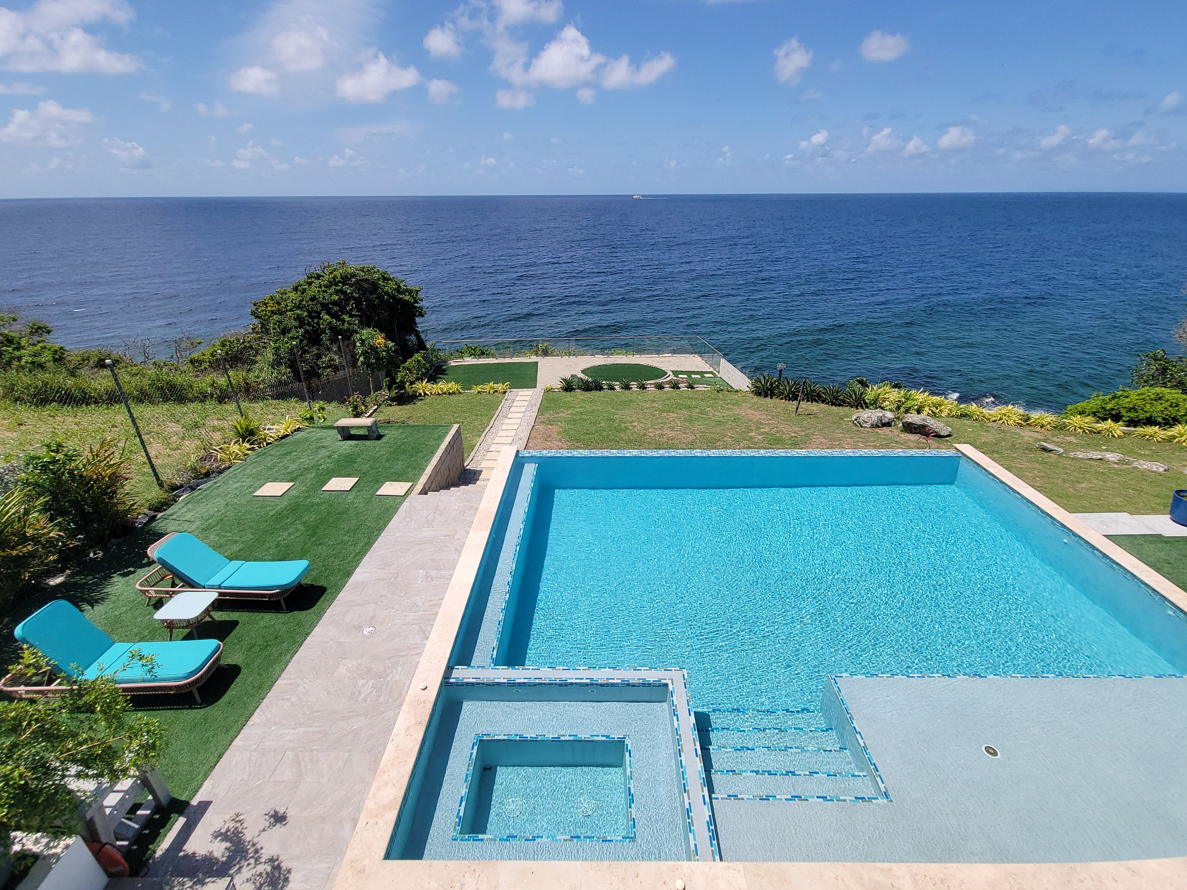 A rectangular swimming pool with a clear blue water feature, surrounded by a paved deck and a small grassy area with a bench.