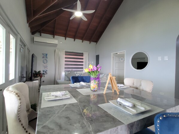 A dining area with a marble table, white upholstered chairs, a ceiling fan, and a view of the adjacent living room.