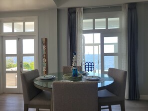 A dining area with a glass table, four chairs, and a view of the sea through large windows.