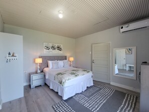 Une chambre à coucher comprenant un lit, deux tables de chevet, une commode, un climatiseur fixé au mur, une porte et un miroir.
