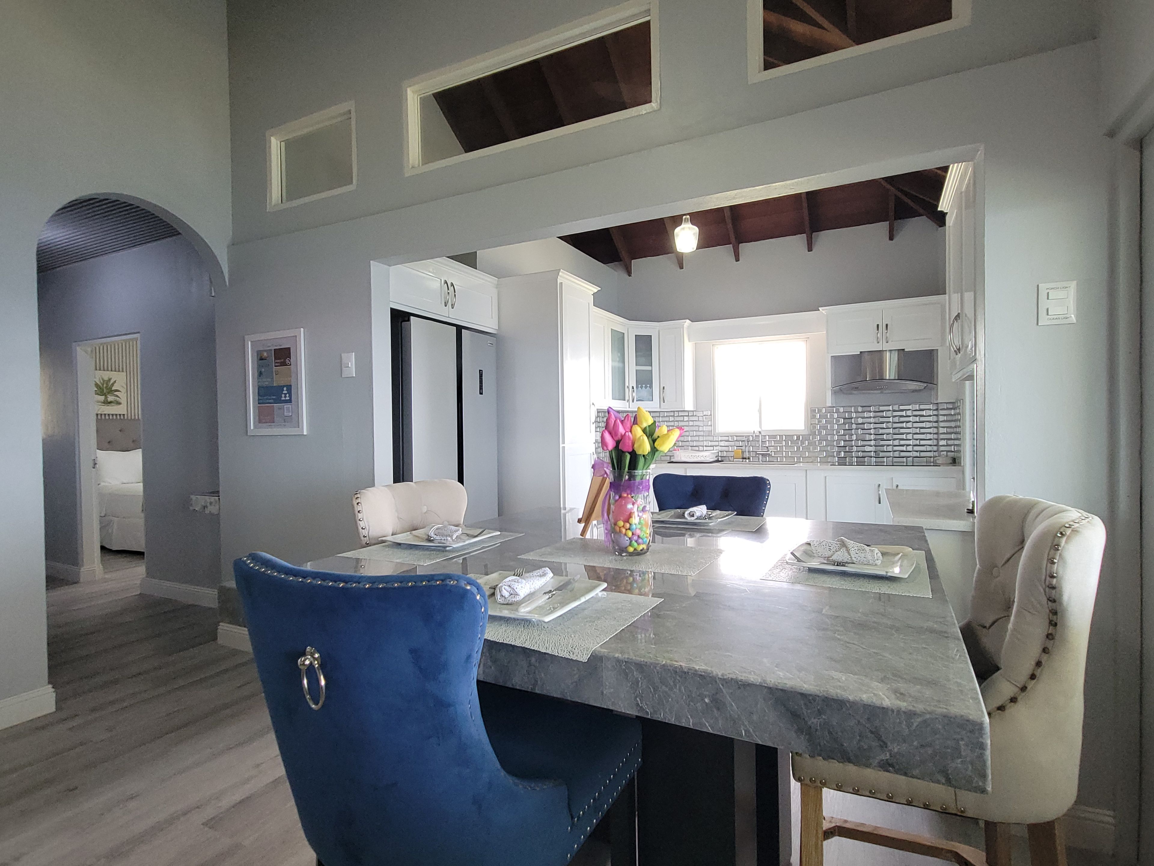 A modern kitchen with white cabinets, a marble countertop, and a dining area with a blue chair and a vase of flowers.