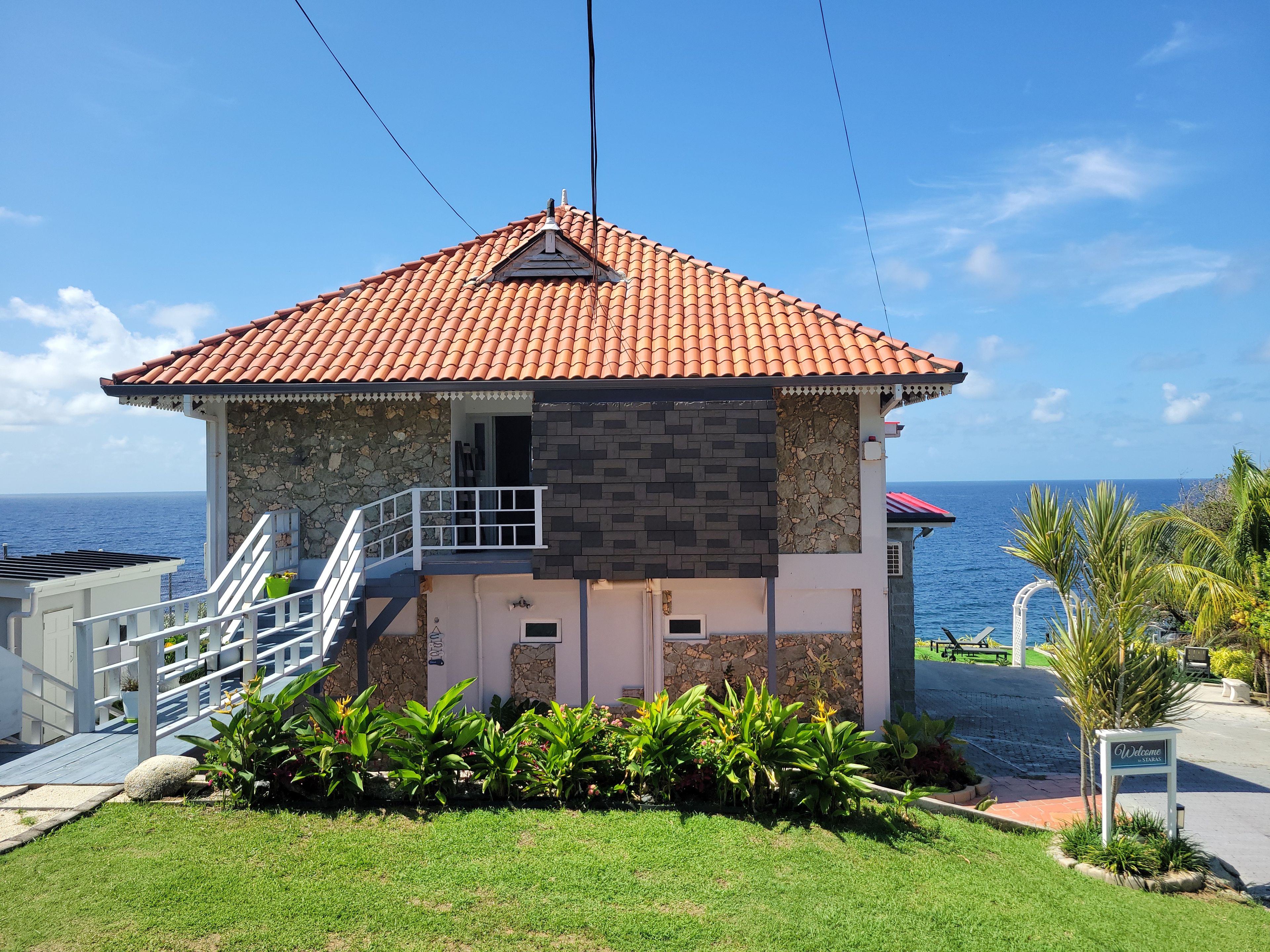 A two-story building with a red-tiled roof, stone and brick walls, a balcony, and a view of the sea.