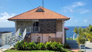 A two-story building with a red-tiled roof, stone and brick walls, a balcony, and a view of the sea.