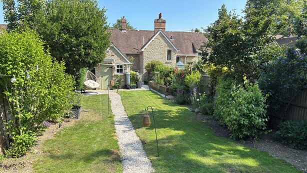 A stone house with a gravel path leading to the front door, surrounded by lush greenery and flowering plants.
