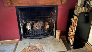 A fireplace with a wooden mantel, a fire, and a stack of firewood to the right.