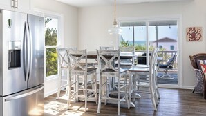 A modern kitchen with a stainless steel refrigerator, a dining table with chairs, and a view of the ocean through large windows. - Bluebird By the Sea: Almost Oceanfront with Private Hot Tub! (Emerald Isle)