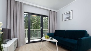 Ein modernes Wohnzimmer mit einem türkisfarbenen Sofa, einem weißen Couchtisch mit Obstschale, einem Flachbildfernseher auf einem weißen Schrank und einer großen Glastür, die auf einen Balkon führt.