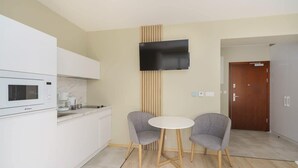 Eine moderne Küche mit weißen Schränken, einer Mikrowelle und einer Kaffeemaschine. Ein Flachbildfernseher ist an der Wand montiert, zwei graue Stühle stehen um einen kleinen runden Tisch, und im Hintergrund ist eine Holztür zu sehen.