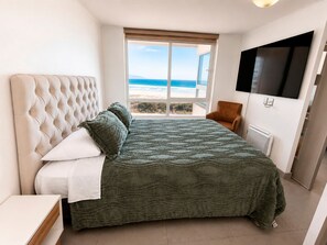 A bedroom with a large bed, a view of the ocean, a television on the wall, and a chair.