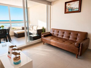A living room with a brown leather sofa, a glass coffee table, and a view of the beach through large sliding doors.