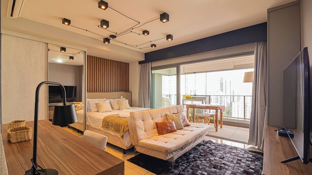 A modern hotel room with a bed, sofa, desk, and a large window offering a city view.