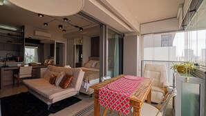 A modern living room with a sofa, a dining table with a red and white tablecloth, a chair, and a balcony with a view of the city.