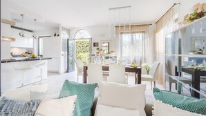 Uma cozinha moderna com armários brancos, bancada de mármore e área de refeições com mesa e cadeiras de vidro.