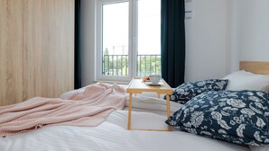 A bedroom with a bed, a bedside table with a cup and saucer, a window with curtains, and a view of greenery outside. - ShortStayPoland Pileckiego B140 (Warsaw)