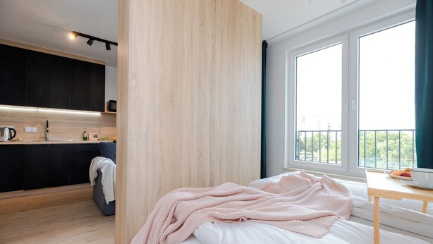A modern hotel room with a bed, a bedside table, a window with a view, a kitchenette, and a wooden partition. - ShortStayPoland Pileckiego B140 (Warsaw)