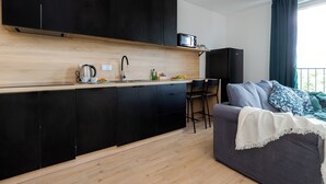 A modern kitchen with black cabinets, a microwave, and a sink. There is a grey sofa with a white throw blanket and blue pillows. - ShortStayPoland Pileckiego B140 (Warsaw)