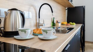 A modern kitchen with a stainless steel kettle, two white cups with green designs, a plate with a croissant, and a fruit bowl. - ShortStayPoland Pileckiego B140 (Warsaw)