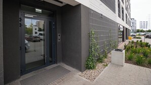 A modern building entrance with a glass door, a metal gate, and a small garden area with plants and flowers. - ShortStayPoland Pileckiego B140 (Warsaw)
