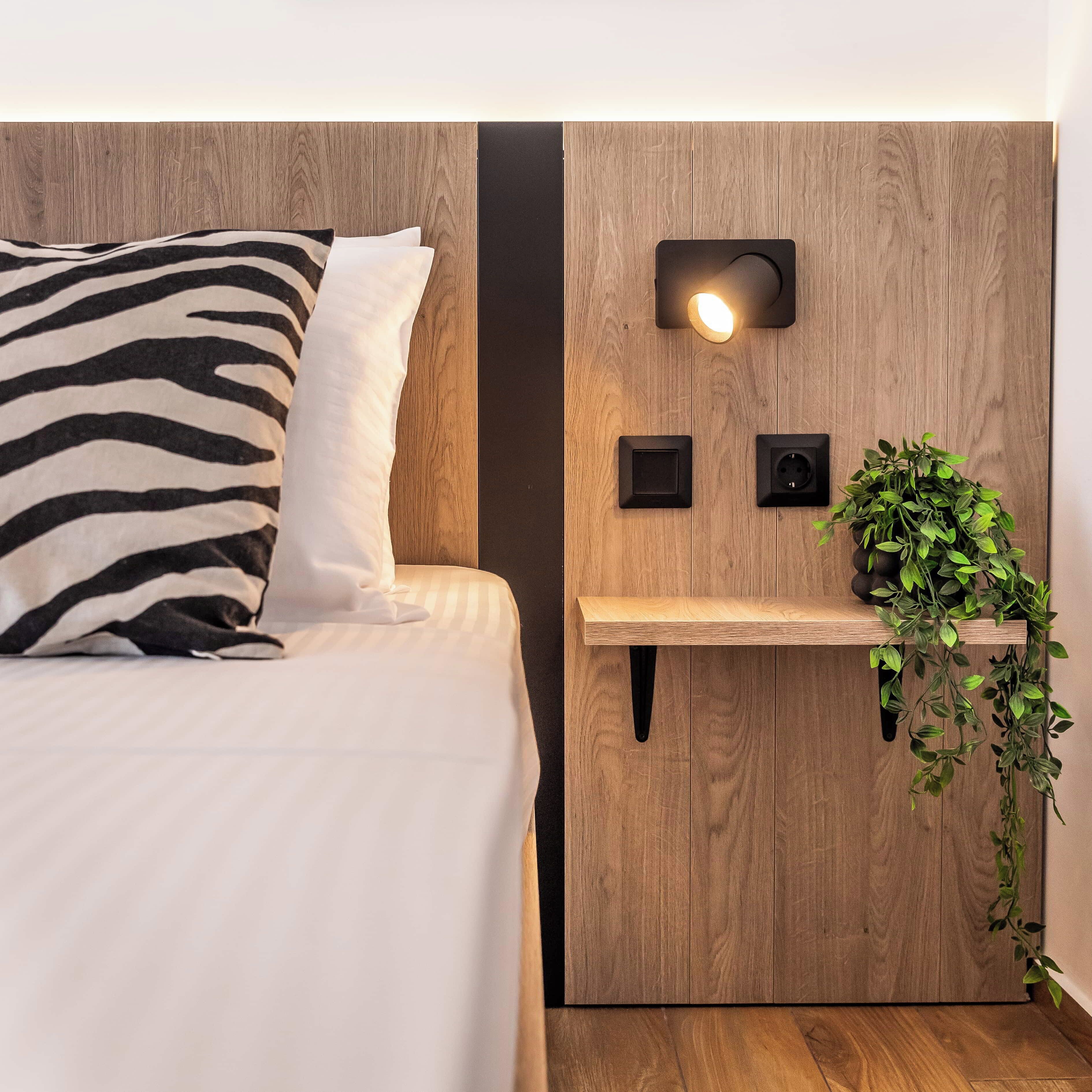A neatly made bed with a zebra-patterned pillow, a bedside table with a wall-mounted light, and a hanging plant.