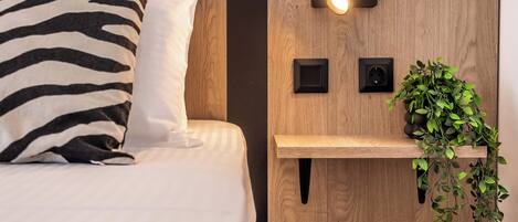 A neatly made bed with a zebra-patterned pillow, a bedside table with a wall-mounted light, and a hanging plant.