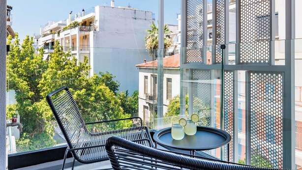 A balcony with a black metal chair, a small round table with two glasses and a bottle, and a view of buildings and trees. - The Kokoon Volos Comfort Living (Volos)