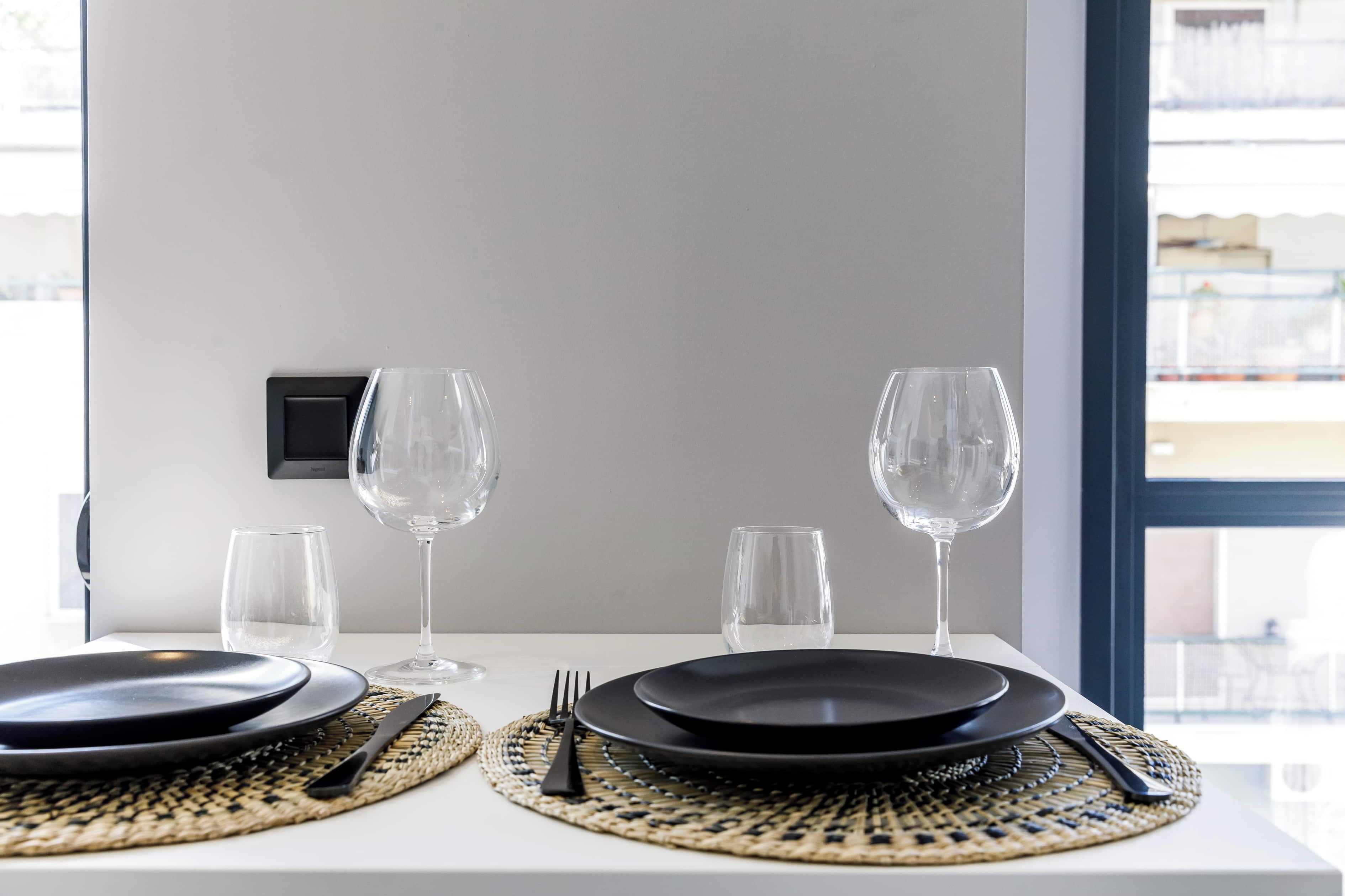 A dining table set with black plates, clear glasses, and cutlery, with woven placemats.