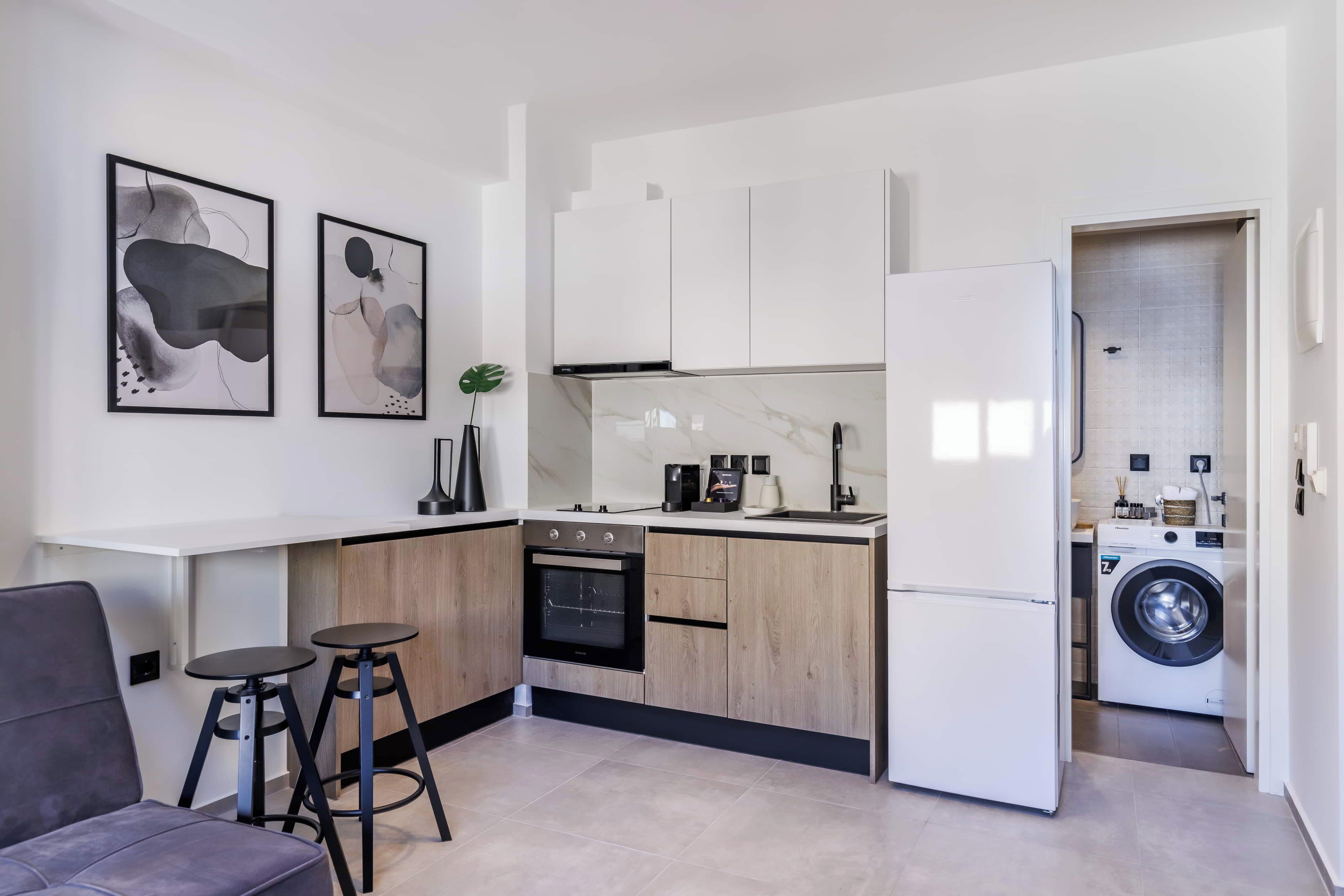A modern kitchen with white cabinets, a built-in oven, a washing machine, and two stools.