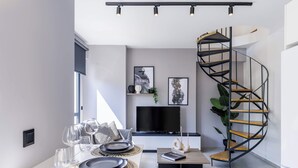 A modern living room with a white dining table set for four, a black TV, and a spiral staircase. - The Kokoon Volos Comfort Living (Volos)