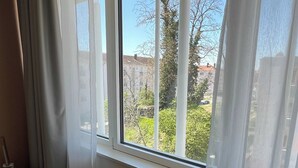 Ein Zimmer mit großem Fenster, weißen Vorhängen und Blick auf Gebäude und Bäume.