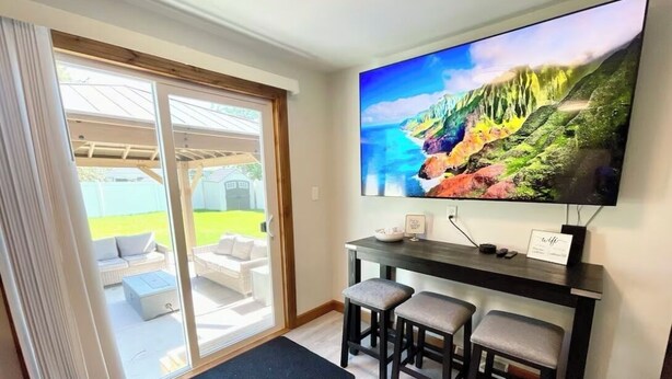 A living room with a large flat-screen TV mounted on the wall, a dark wooden coffee table, and three bar stools. - Great Location close to Resch/Lambeau, Sleeps 14, Huge Game Room, Private Yard (Green Bay)