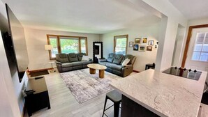 A modern living room with a sofa, coffee table, and a kitchen area with a marble countertop and bar stools. - Great Location close to Resch/Lambeau, Sleeps 14, Huge Game Room, Private Yard (Green Bay)