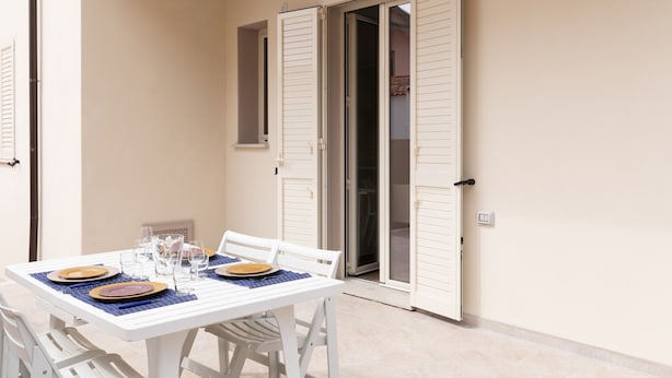 A white table set with plates, glasses, and a placemat on a balcony with white shutters and a light fixture above. - Casa Marina con Patio by Wonderful Italy (Olbia)