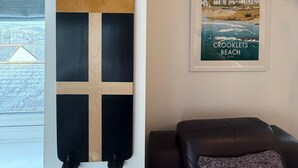 A room with a large window, a black skateboard, a framed poster of Crooklets Beach, and a black leather sofa.