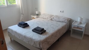 Un lit bien fait, recouvert d’une couvre-lit à motifs de feuilles, deux serviettes pliées, une table de chevet avec une lampe et une prise électrique au mur.