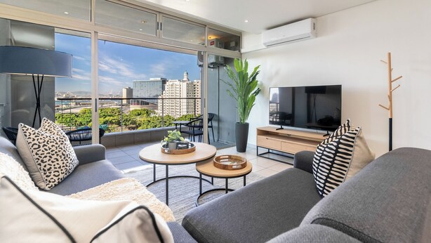 A modern living room with a large TV, comfortable seating, a coffee table, and a view of the cityscape.