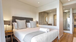 A modern hotel room with a large bed, a wooden nightstand, a mirror, and a bathroom visible through an open door.