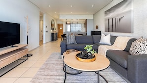 A modern living room with a flat-screen TV, a sofa, a coffee table, and a dining area in the background.