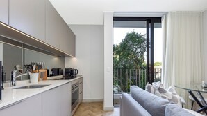 A modern kitchen with a built-in oven and sink, a grey sofa, and a balcony with a view of trees.