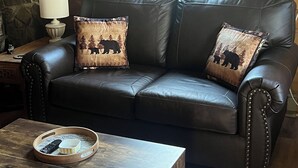 Een donkerbruine leren bank met twee decoratieve kussens, een houten salontafel met dienblad en afstandsbediening, en een stenen muur met een ingelijst schilderij.