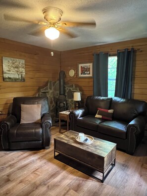 A cozy living room with a stone fireplace, leather armchairs, a wooden coffee table, and a ceiling fan.