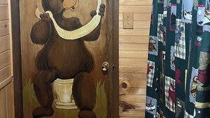 Een houten deur met een tekening van een beer die op een toilet zit, een quiltgordijn met verschillende patronen, en een houten muur.