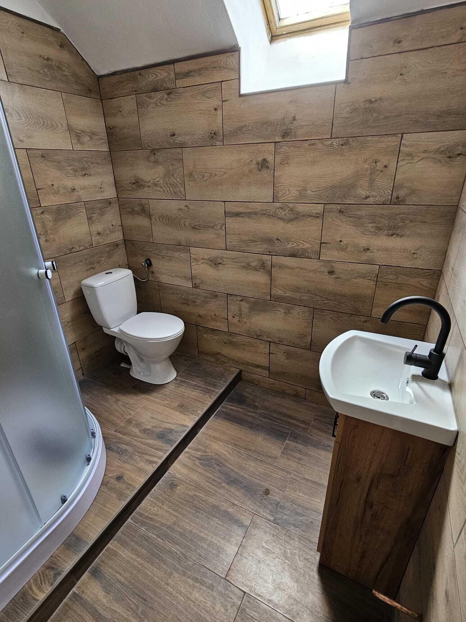 A small bathroom with wooden wall tiles, a toilet, a sink, and a skylight.