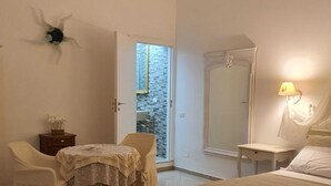 A hotel room with a bed, a table with a chair, a small bedside table, a lamp, and a view through a door.