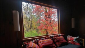 A room with a large window overlooking a forest with autumn foliage. There is a bed with multiple pillows and a bedside table. - Cozy, welcoming Berkshires cottage – perfect home base & place to unplug! (Stockbridge)