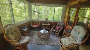 A cozy room with wicker chairs, a hanging chair, a small table, and a view of the outdoors. - Cozy, welcoming Berkshires cottage – perfect home base & place to unplug! (Stockbridge)