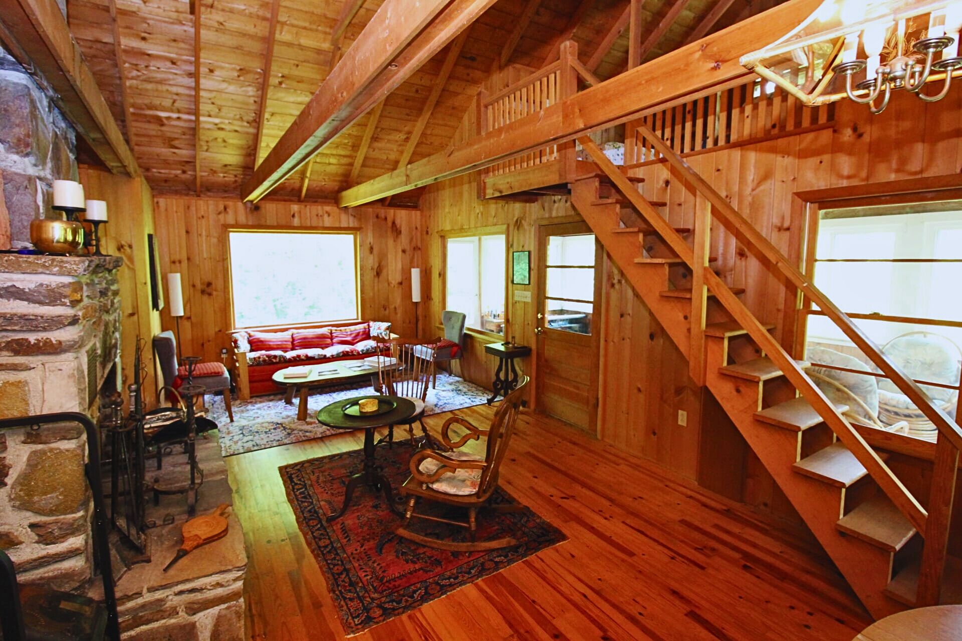 Interior acogedor de una cabaña con chimenea de piedra, paredes de madera, una escalera y un ventanal grande.